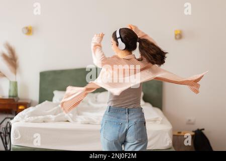 Happy young Caucasian woman wearing headphones spinning dance and enjoy life. Back view. Leisure in home. Stock Photo