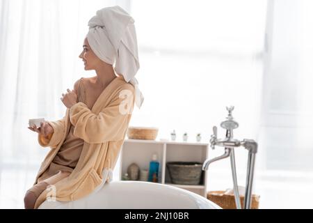 Side view of young woman in bathrobe and towel holding cosmetic cream near bathtub in bathroom,stock image Stock Photo