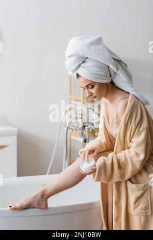 Young woman in towel and bathrobe holding body cream near bathtub at home,stock image Stock Photo