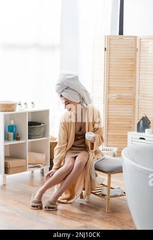 Young woman in towel and bathrobe applying body cream on leg in bathroom,stock image Stock Photo