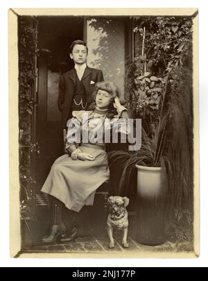 Original Victorian era photograph of stroppy / belligerent looking teenagers - a middle class attractive boy and girl, probably brother and sister. A small pug dog sits next to them, wearing a collar with bells on it - a naughty one! It's a relaxed, informal photograph taken outside a house in the doorway of their home, potted plants next to them, circa 1899 Stock Photo