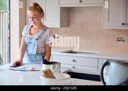 Woman Renovating Kitchen At Home With Paint Swatches Using Mobile Phone Stock Photo