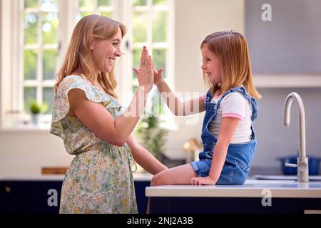 Family At Home With Daughter Sitting On Kitchen Counter Giving Mother High Five Stock Photo