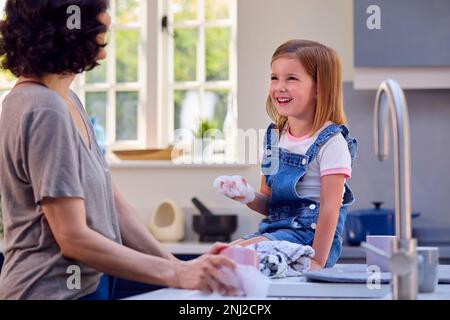 Family With Daughter Helping Older Mother To Do Washing Up In Kitchen At Home Blowing Soap Bubbles Stock Photo