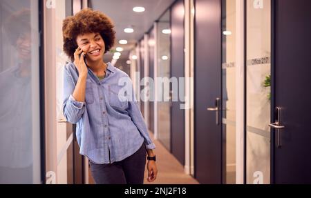 Smiling Young Businesswoman In Corridor Of Modern Office Talking On Phone Stock Photo