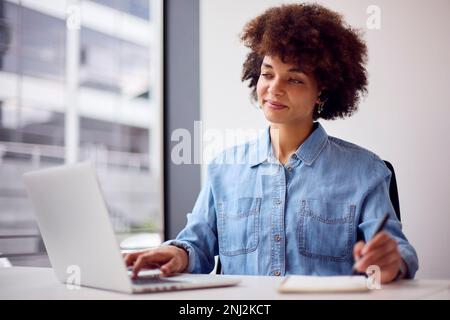 Young Businesswoman In Modern Office Working On Laptop Using Wireless Earpiece Stock Photo
