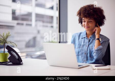 Young Businesswoman In Modern Office Working On Laptop Using Wireless Earpiece Stock Photo