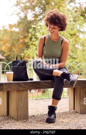 Young Woman Outdoors In Park Sitting On Bench Working Writing In Notebook Stock Photo