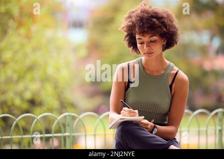Young Woman Outdoors In Park Sitting On Bench Working Writing In Notebook Stock Photo