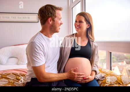 Loving Couple At Home Sitting On Bed With Man Touching Pregnant Woman's Stomach Stock Photo