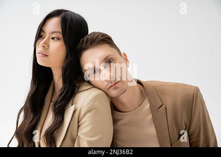 stylish man in beige blazer leaning on young asian woman with long brunette hair isolated on grey,stock image Stock Photo