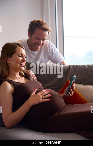 Couple With Pregnant Woman Holding Mobile Phone Relaxing On Sofa At Home Holding Stomach Stock Photo