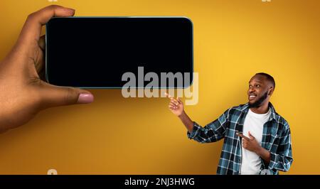 Positive Black Man Pointing Aside At Huge Female Hand With Blank Smartphone Stock Photo