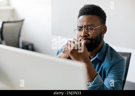 Thoughtful black man in eyeglasses stack with hard task, looking at computer screen, thinking of problem solution Stock Photo