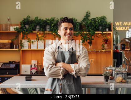 Confident barista in an apron. Young guy working as a barista in a coffee shop standing with crossed hands and looking away. Stock Photo