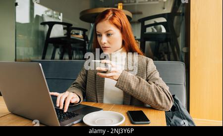 Woman working as a digital nomad in a cafe. Female entrepreneur with ginger hair holding a cup typing on a laptop Stock Photo