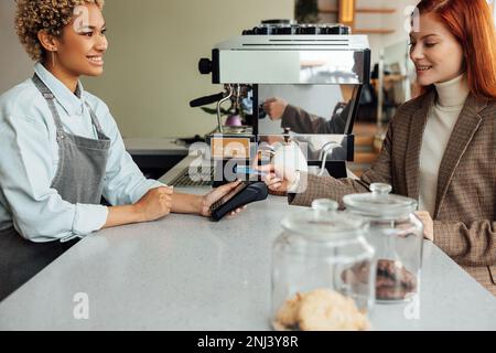 Client paying by card at a coffee shop. Smiling barista holding pos terminal while customer paying. Stock Photo