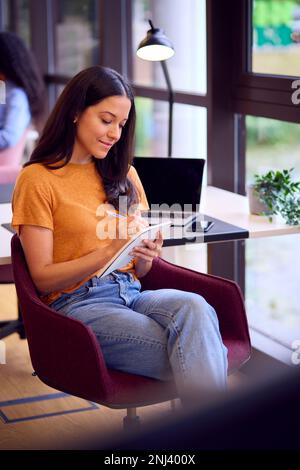 Businesswoman Sitting At Desk In Office Making Notes In Notebook Stock Photo