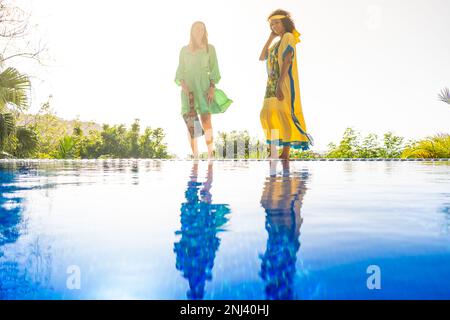 Two women with colorful dress posing next to an outdoor pool with the image reflected on the water Stock Photo