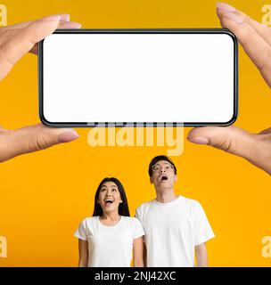 Amazed Asian Couple Looking Up At Big Smartphone In Huge Hands Stock Photo