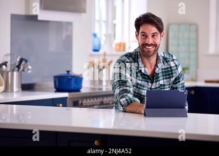 Man Using Digital Tablet In Kitchen To Work From Home Book Holiday Or Shop Stock Photo