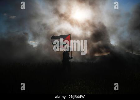 Gaza City. Palestine. February 22nd, 2023. A demonstrator holds the Palestinian flag in front of burning tires during a demonstration along the border between the Gaza Strip and Israel, in solidarity with Nablus city against the Israeli military operations that killed at least ten Palestinians. Stock Photo
