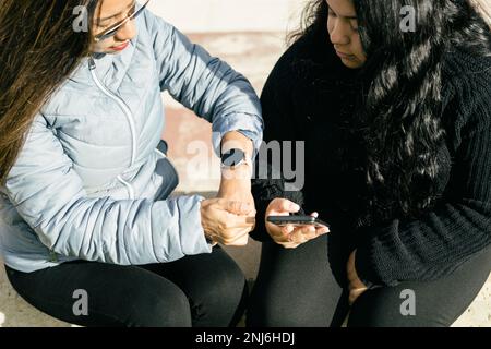 Hispanic mother in sunglasses checking time on smartwatch while sitting on bench with daughter using smartphone in park on sunny day Stock Photo