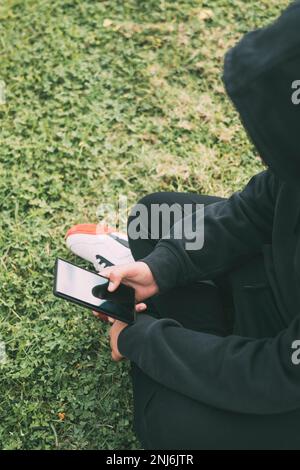young man sitting on the ground in the park, completely immersed in his smartphone. Stock Photo