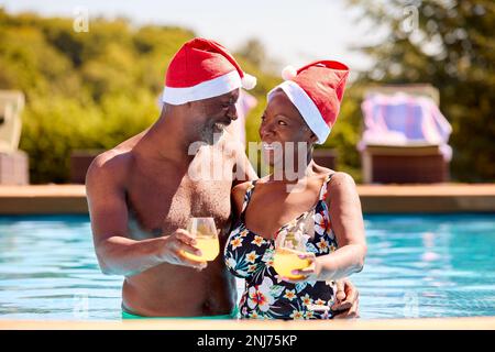 Senior Couple On Christmas Holiday In Swimming Pool Wearing Santa Hats Stock Photo