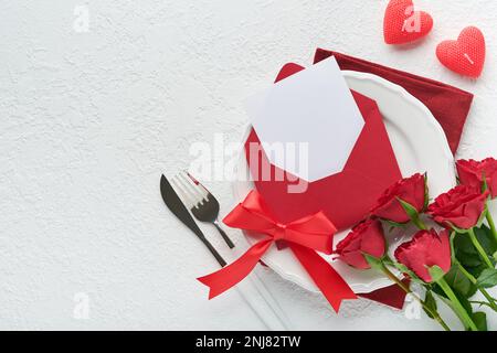 Romantic creative table setting. Empty white plate and red roses, candles, knife, fork and decorative silk hearts on white background. Valentines Wedd Stock Photo