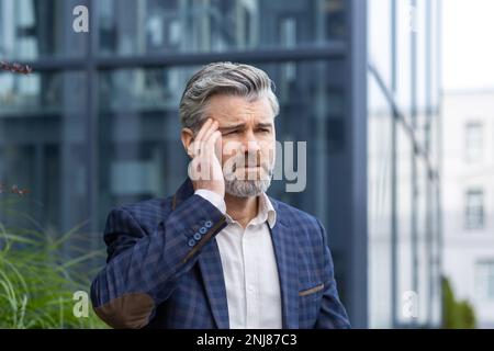 Mature fair-haired man outside office building has dizziness and headache, businessman in business suit is overwhelmed in the air, senior boss is sick. Stock Photo