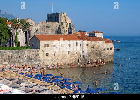 Pebble beach with sunbathers in summer outside the Venetian city walls of Budua at the medieval town Budva along the Adriatic Sea, Montenegro Stock Photo