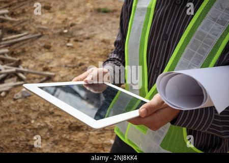 Asian business man construction manager and engineer worker in protective helmet hold blueprints paper on hand using tablet computer at building site Stock Photo