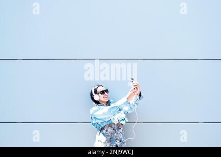 Young Latina woman with pink headphones and black glasses with her arms raised taking a photo on a gray background in summer Stock Photo