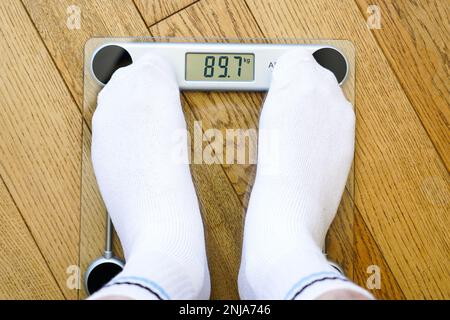 A man in white socks weighing himself on a transparent electronic floor scale, the weight reading is 89 kg, top view Stock Photo