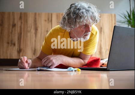 Authentic senior man lying on the living room floor looking at laptop computer and making notes in a notepad. Active and inquisitive retired life. Stock Photo