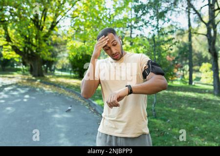 Young African American man runner, athlete in the park. A tired man counts his pulse on a fitness bracelet. Surprised, he looks at the result on the smartwatch on his hand. Wipes sweat with his hand. Stock Photo