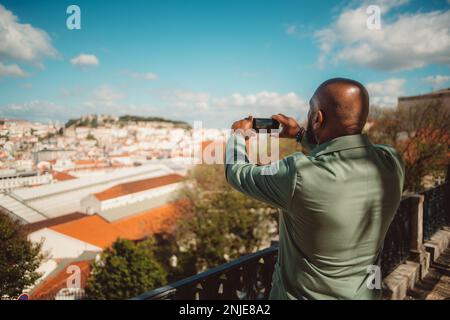 Capture in selective focus of a bald, bearded black tourist male from behind in a green suit jacket takes pictures of the city from a viewpoint on his Stock Photo