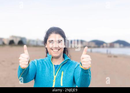 Hispanic woman standing doing happy thumbs up gesture with hand. approving expression looking at the camera showing success. Stock Photo