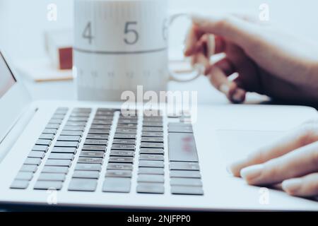 in-focus detail of a laptop computer keyboard, next to a heavily manicured female hand near the touchpad, and in the background a mug with a hot drink Stock Photo