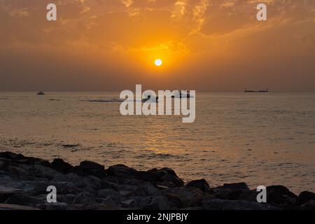 Sunset at the sea, the sun close to the sea horizon, the whole cloudy sky shines in intense gold orange colors, on the sea are boats. Stock Photo