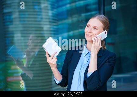 Smart professional caucasian business woman uses mobile phone in city outdoors holding documents in her hands, dressed in a blue shirt on the background of a business center Stock Photo