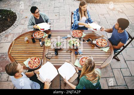 Time to feast. High angle shot of a group of friends sitting around a table and enjoying pizza together outdoors. Stock Photo