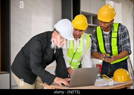 Professional Caucasian businessman in white hardhat and business suit using laptop while meeting and discussing the building plan with a team of const Stock Photo