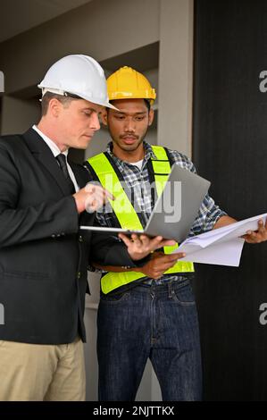 Determined Asian male foreman or construction engineer looking at laptop screen, checking scale on blueprint, discussing working with a Caucasian busi Stock Photo