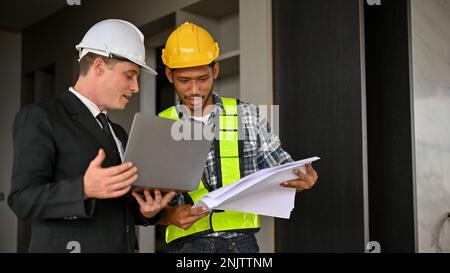 Determined Asian male construction engineer checking scale on blueprint and discussing working with a Caucasian businessman while inspecting the build Stock Photo