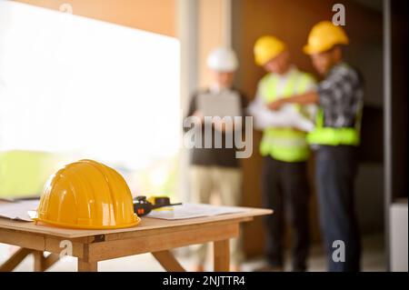 A yellow hardhat, a walkie-talkie, a measuring tape and a blueprint on a table over blurred background of a group of construction workers discussing w Stock Photo