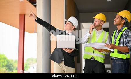 Successful Caucasian businessman in a white hardhat and business suit inspects the building at the site with a construction team. Diverse workers conc Stock Photo