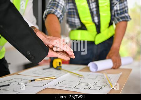Cropped image of a team of construction workers and a businessman putting their hands together over the working table, showing spirit, teamwork. Stock Photo