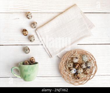 Craft creative Easter layout. Quail eggs on woven plate and falling to green cup and cotton napkin with space for text on white wooden background. Top Stock Photo